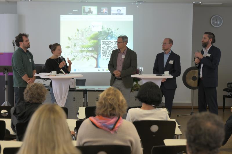 fünf Personen stehen in einem Hörsaal an zwei Stehtischen und diskutieren