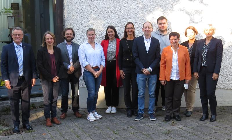 11 Personen vor einer weißen Gebäudewand. Sie stehen in einer Reihe